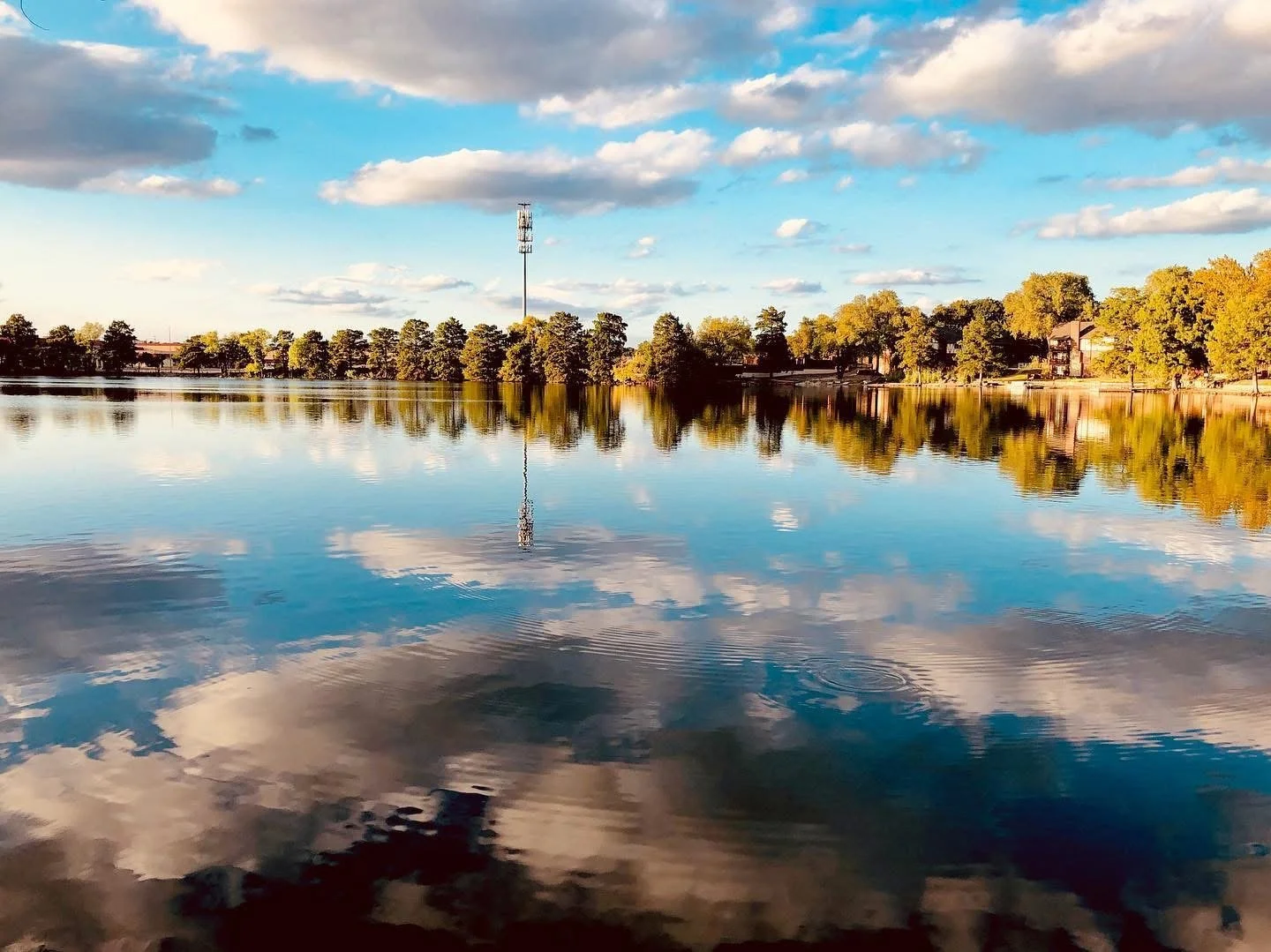 A calm lake reflecting the blue sky, white clouds, and trees along the shoreline at sunset