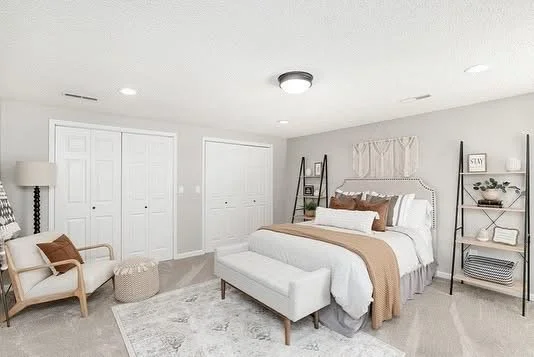 A modern bedroom with a white bed, beige bedding, and decorative pillows, flanked by black metal shelves with books and decor, and a seating area with an armchair and ottoman.