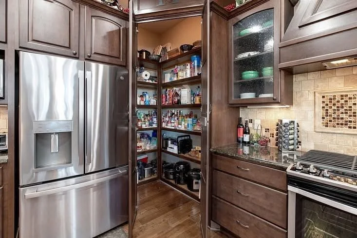 A kitchen with dark wood cabinets, a stainless steel refrigerator, and a pantry filled with various food and kitchen items.
