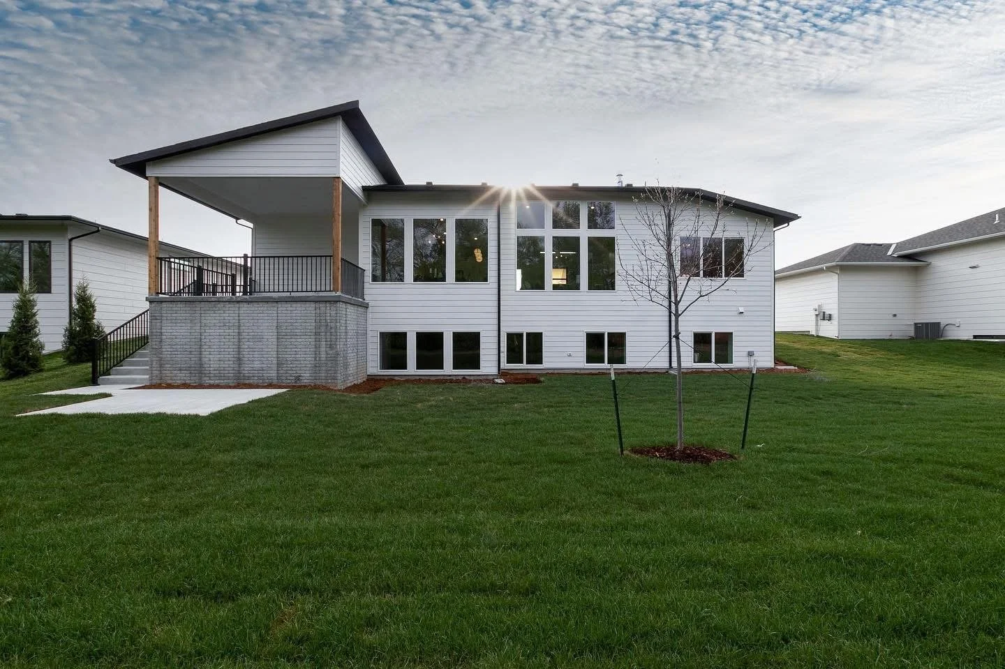 Back view of a modern white house with large windows, a small balcony, and a landscaped backyard with a young tree, green grass, and neighboring houses in a suburban setting under a partly cloudy sky.