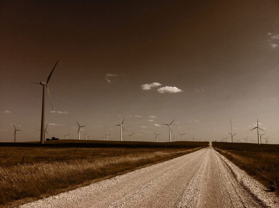 Gravel road leading towards wind turbines in an open field during daytime with a partly cloudy sky.