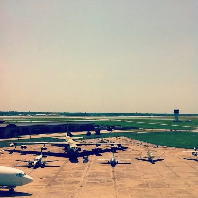Multiple military aircraft parked on tarmac at an airport, with control towers and runways in the background.
