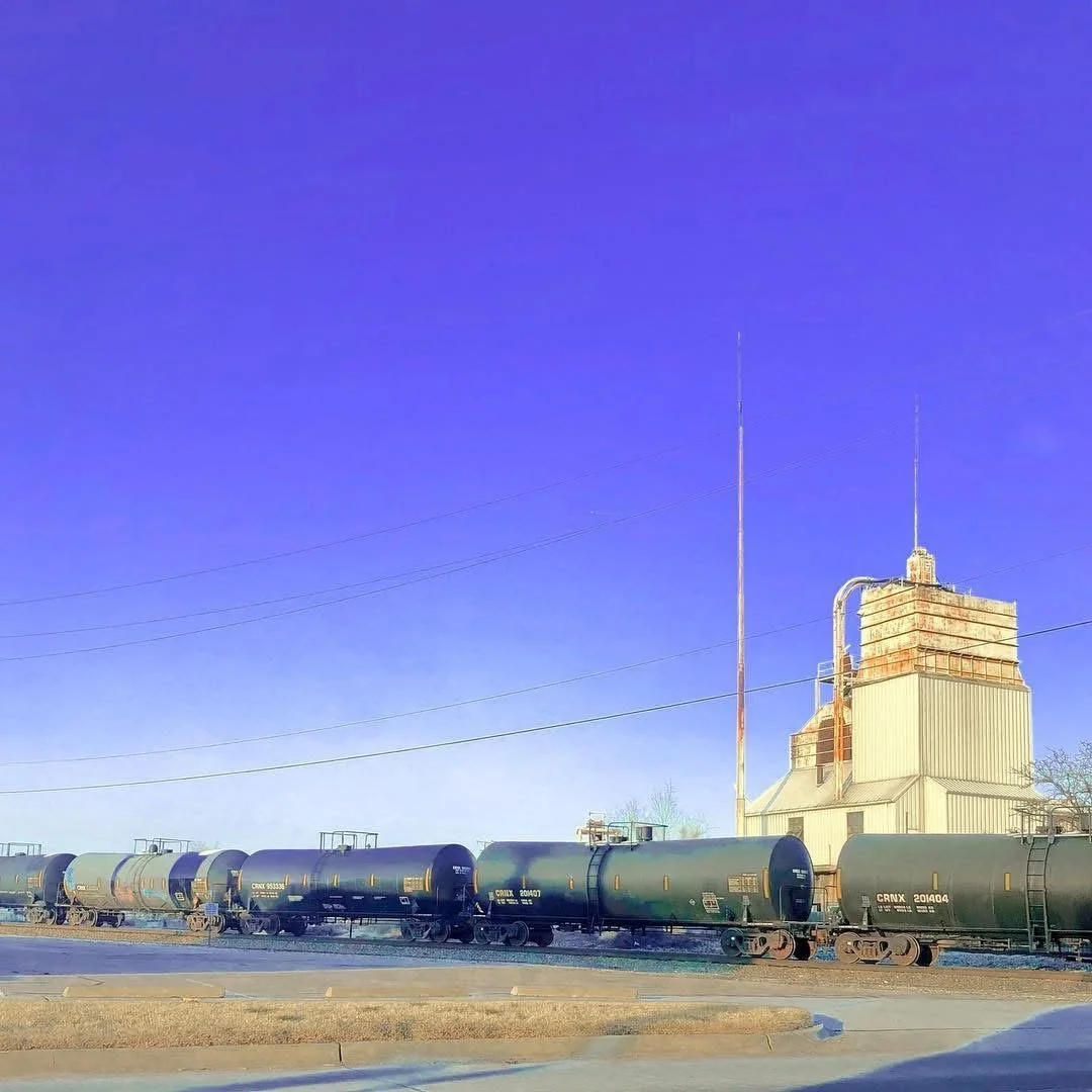 A row of black tanker train cars on tracks with a beige industrial building with rusted metal parts and two tall antennas against a blue sky.