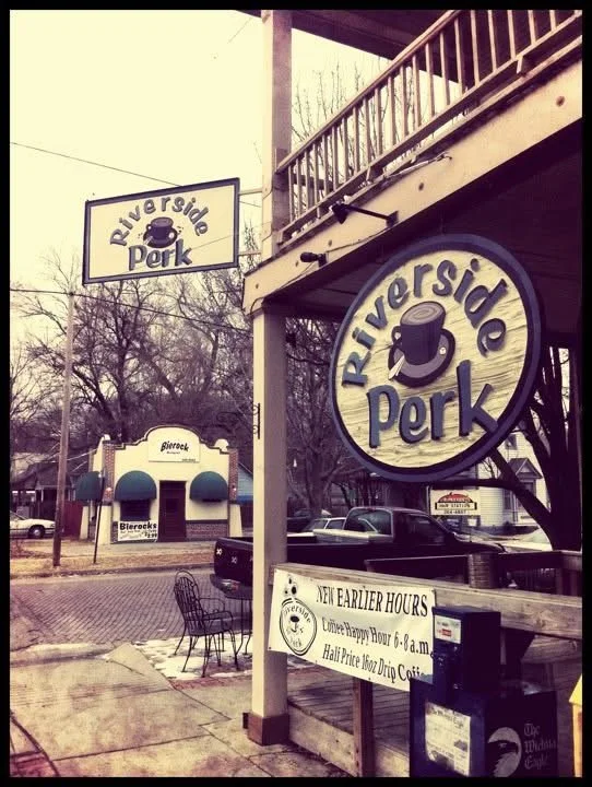 Riverside Park sign with a coffee cup logo, storefront with a large circular Riverside Park sign, outdoor seating, and a sign indicating new hours from early morning.