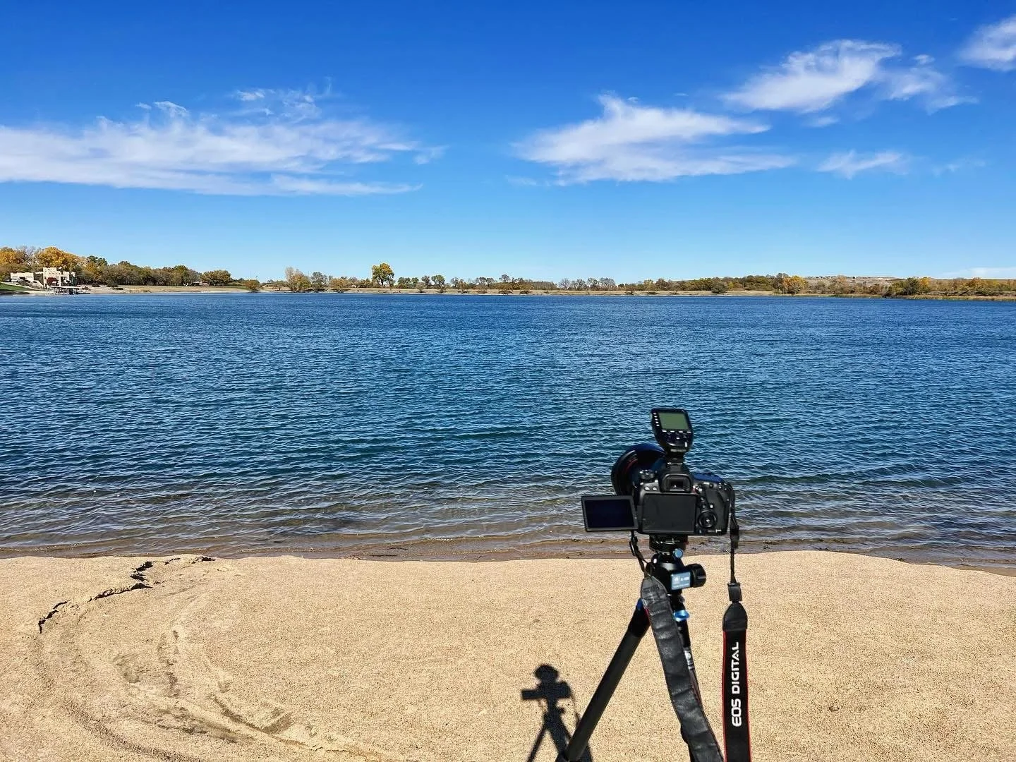 A camera set up on a tripod on a sandy beach facing a lake with blue water and a few clouds in the sky.