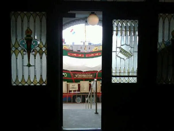 View through an open door with stained glass windows on either side, showing a boat with a white canopy and decorations, and a sign that reads 'WINDWAGON' on a pier or dock.