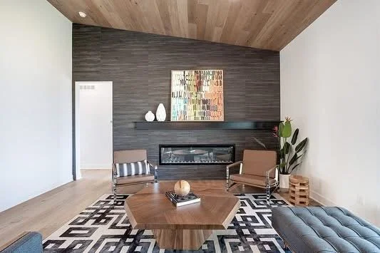 Modern living room with a wood ceiling, gray textured wall, fireplace, abstract artwork, striped armchairs, potted plant, wooden side table, and a patterned black and white rug.