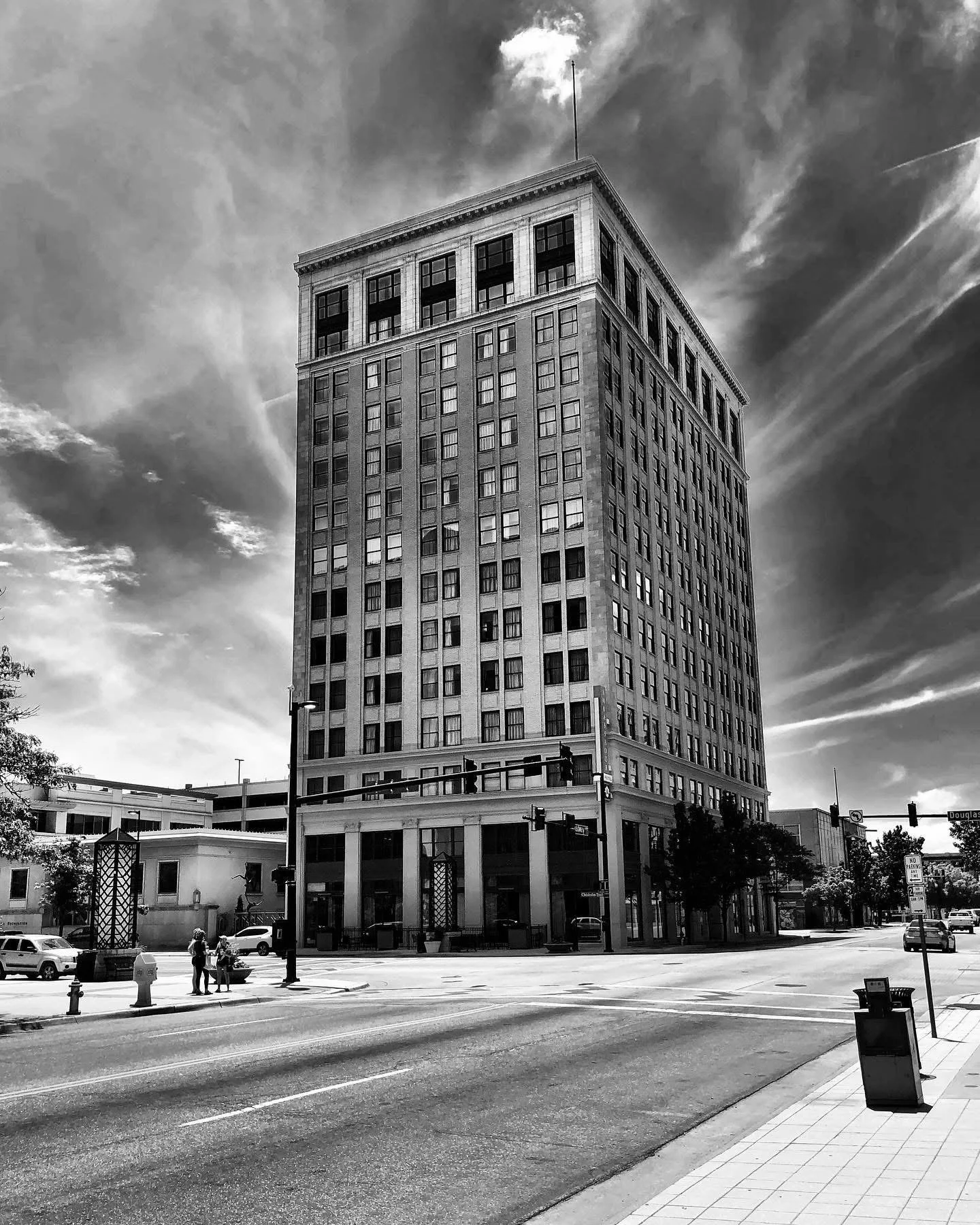 Black and white photo of a tall building on a city street corner with a few people and cars at the intersection.