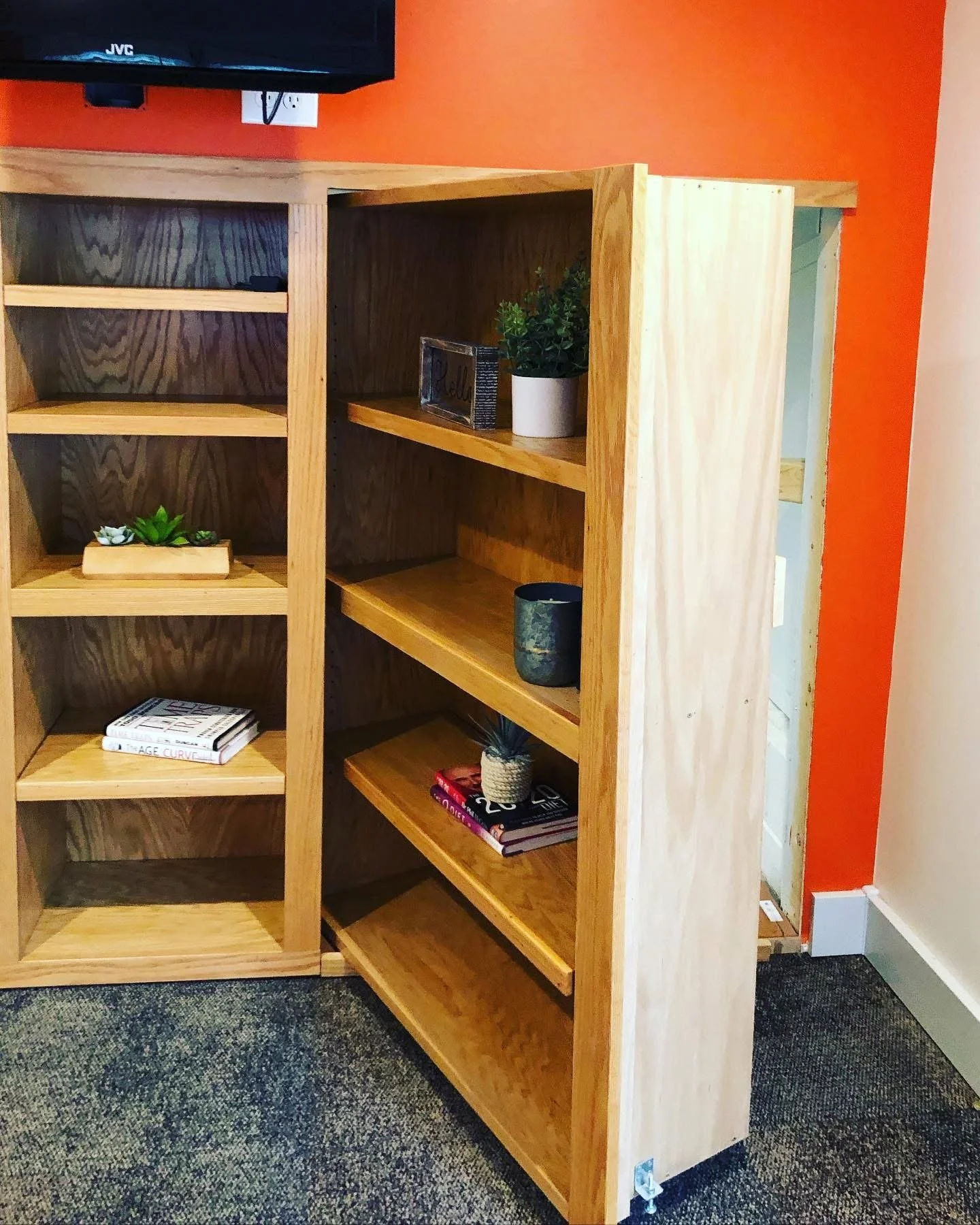 Wooden bookshelf with natural finish, positioned near an orange wall, containing potted plants, books, and decorative items.