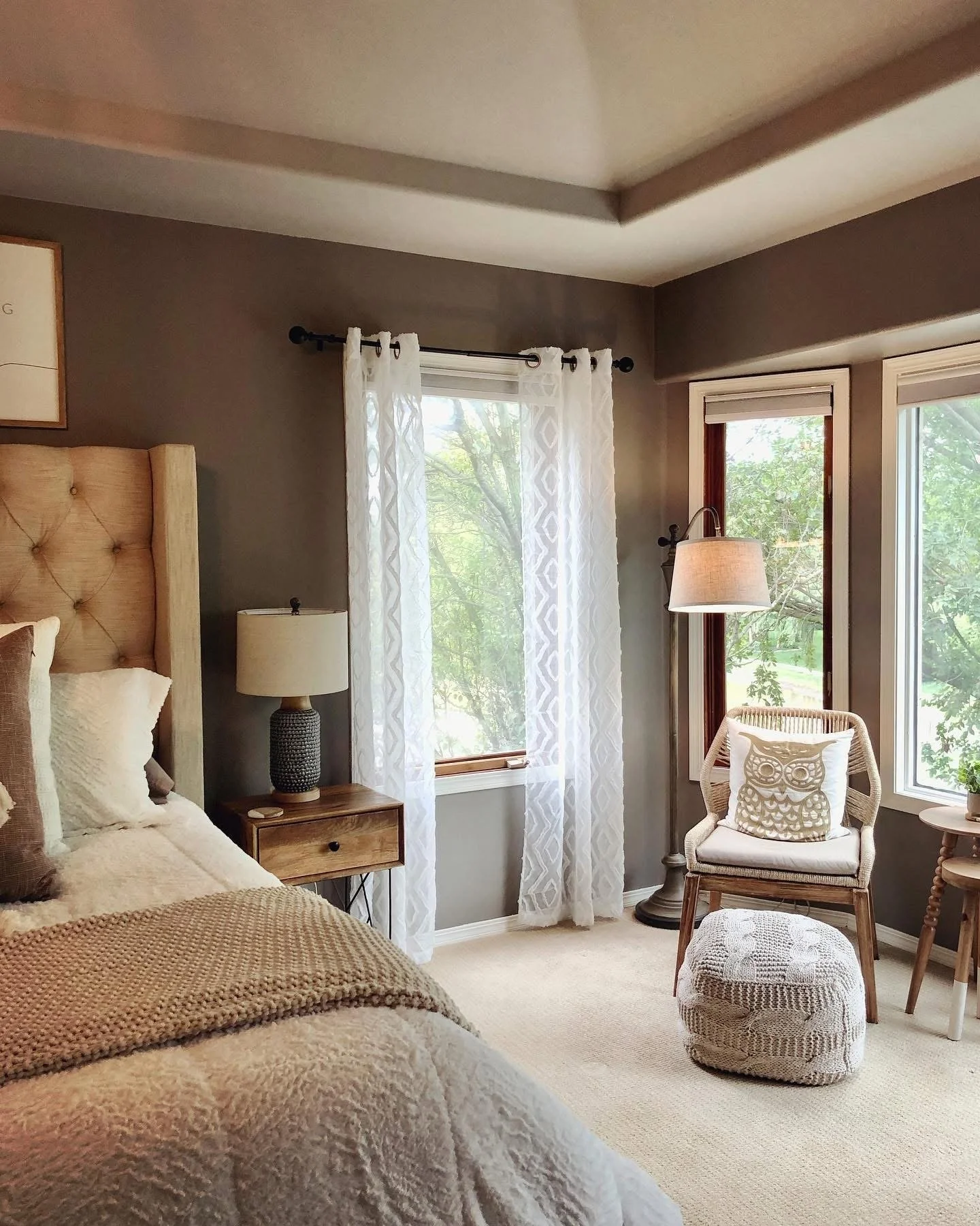 A cozy bedroom with a bed, bedside table, and lamp. There are two windows with sheer white curtains and a chair with an owl pillow, next to a small round table with a plant.