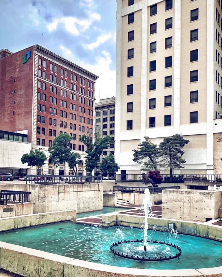 Cityscape with fountain in foreground, high-rise buildings and trees in background, overcast sky.