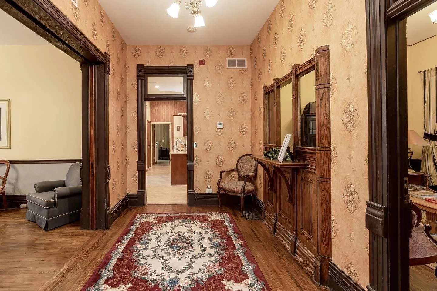 View of a vintage-style foyer with wooden trim, patterned wallpaper, and a floral area rug, leading to a hallway and other rooms in a house.
