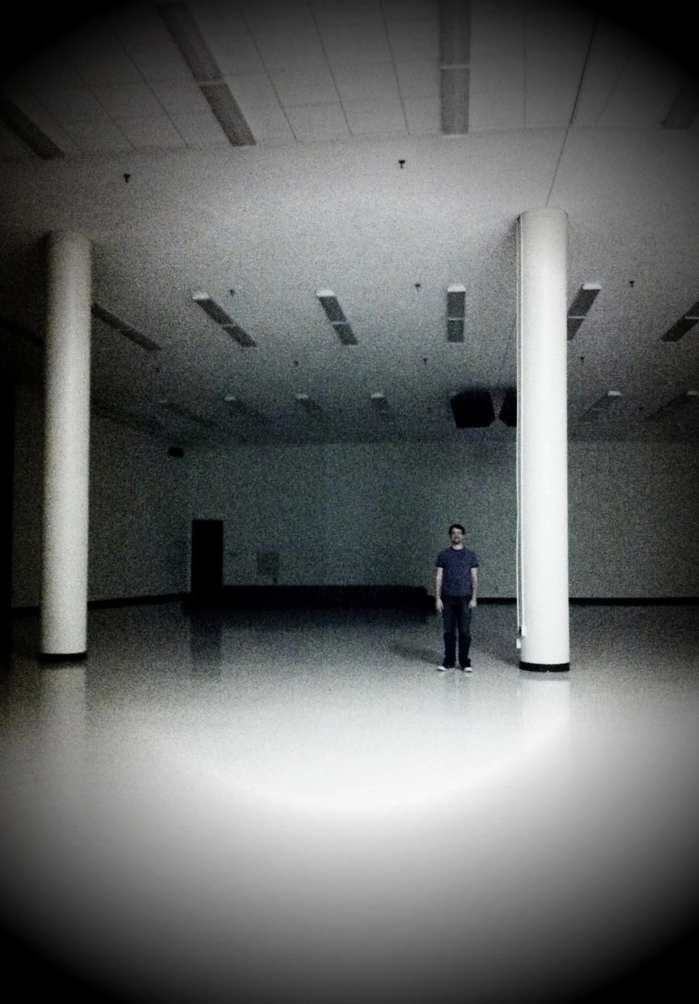 A person standing alone in a large, empty, dark room with two large white columns and a high ceiling with rectangular light fixtures.