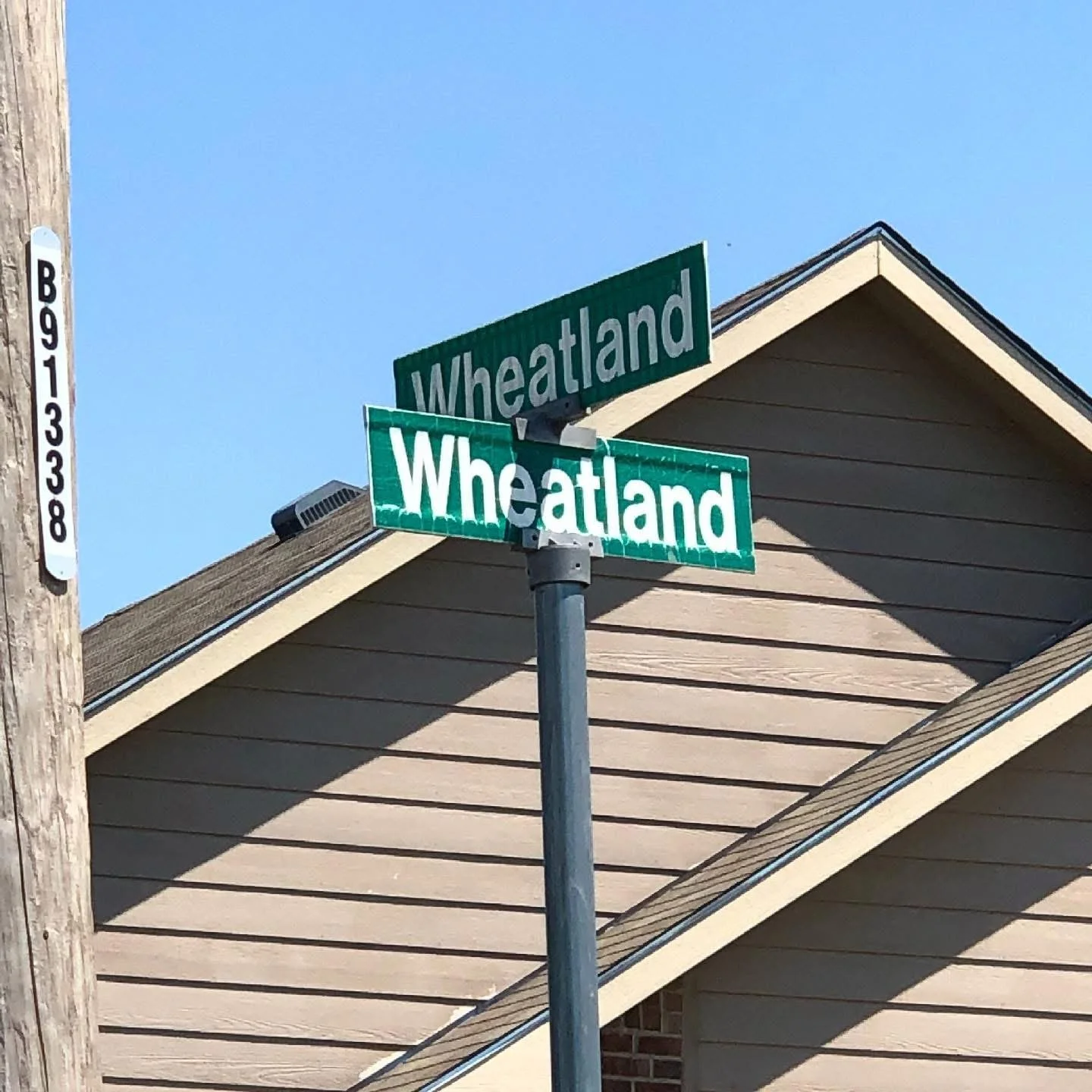 Street signs showing the intersection of Wheatland Avenue. A house with beige siding and a gabled roof is in the background, along with a wooden utility pole with a vertical white and black number plate.