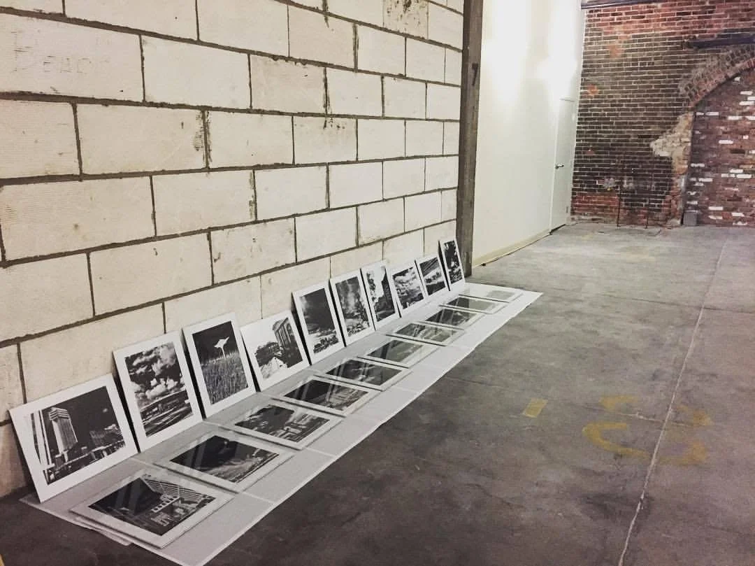 Black and white photographs of cityscapes and buildings are arranged on the floor against a brick wall in an industrial-style space.