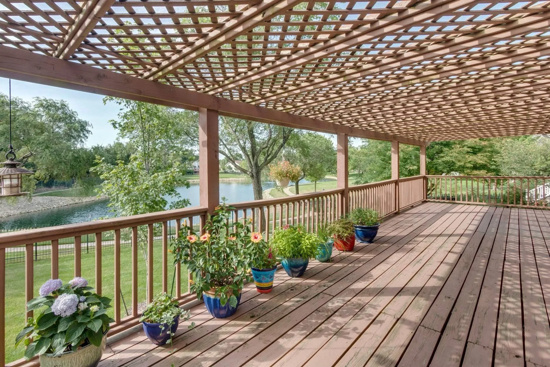 A spacious wooden balcony with a lattice roof, decorated with colorful potted plants along the railing. The balcony overlooks a serene pond surrounded by lush green trees and a well-kept lawn.