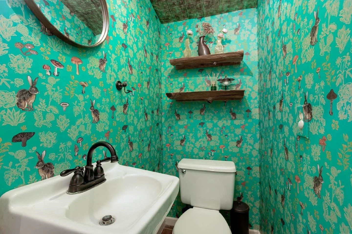 A small bathroom with green wallpaper decorated with rabbits, mushrooms, and plants. It contains a white sink with black fixtures, a toilet, two rustic wooden shelves with decorative vases and flowers, and a round mirror.