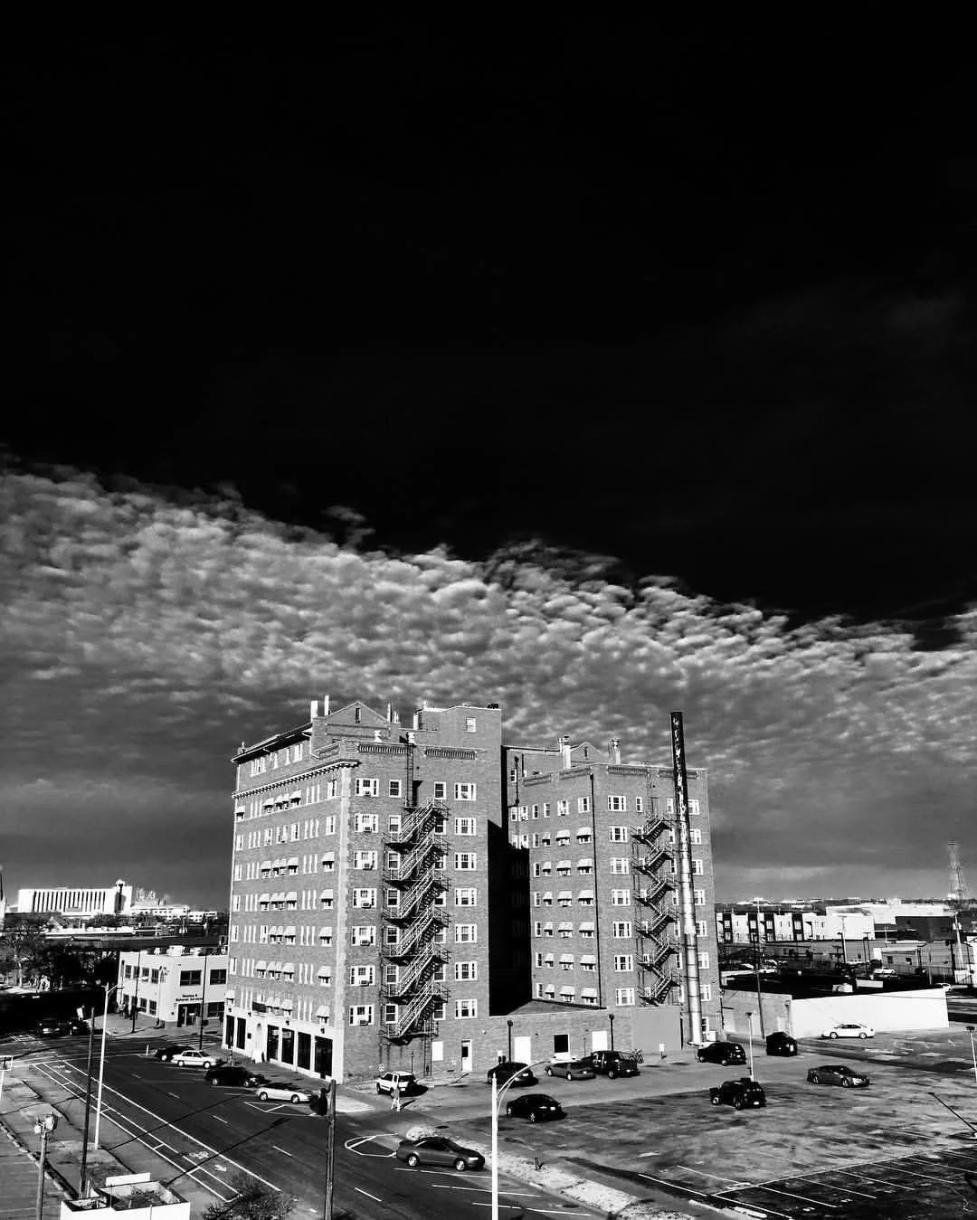 Black and white photo of a multi-story building with fire escape stairs and surrounding parking lot, under a cloudy sky.