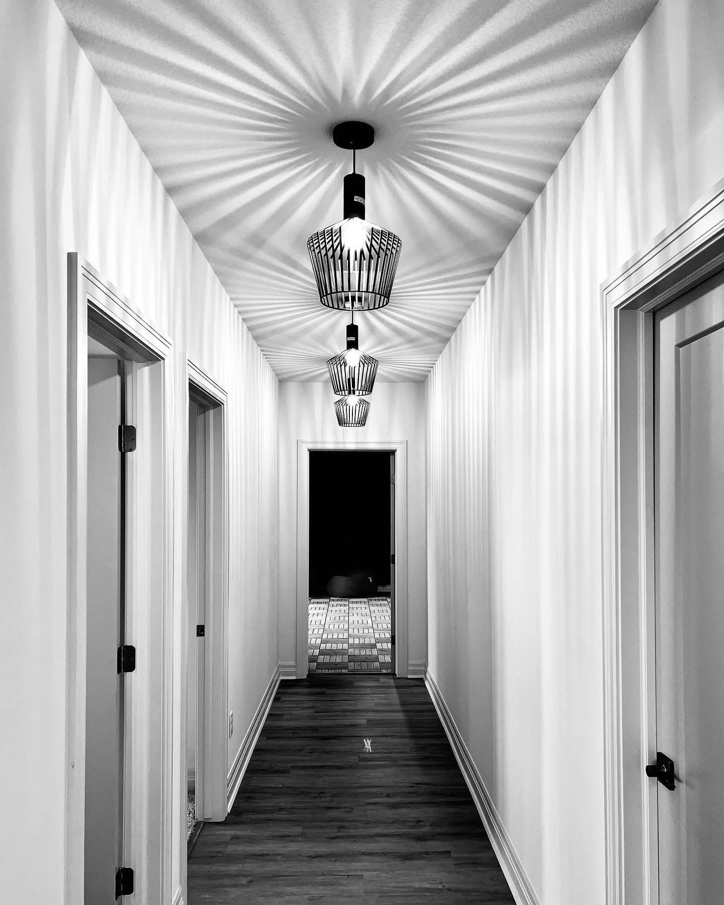 Black and white photo of a hallway with three pendant lights casting starburst shadows on the ceiling. The hallway has white paneling walls, wood flooring, and doorways on each side leading to different rooms. At the end of the hallway, there is a do