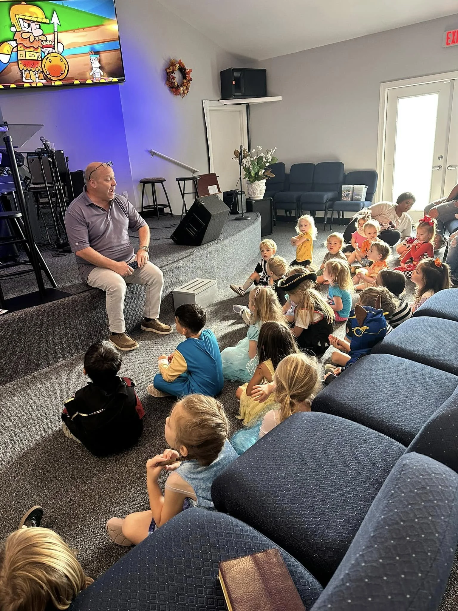 A man sitting on a small stage with children sitting on the carpeted floor, watching him. The children are dressed in costumes, and a TV screen showing cartoon characters is visible in the upper left corner. The scene is inside a room with a door, so
