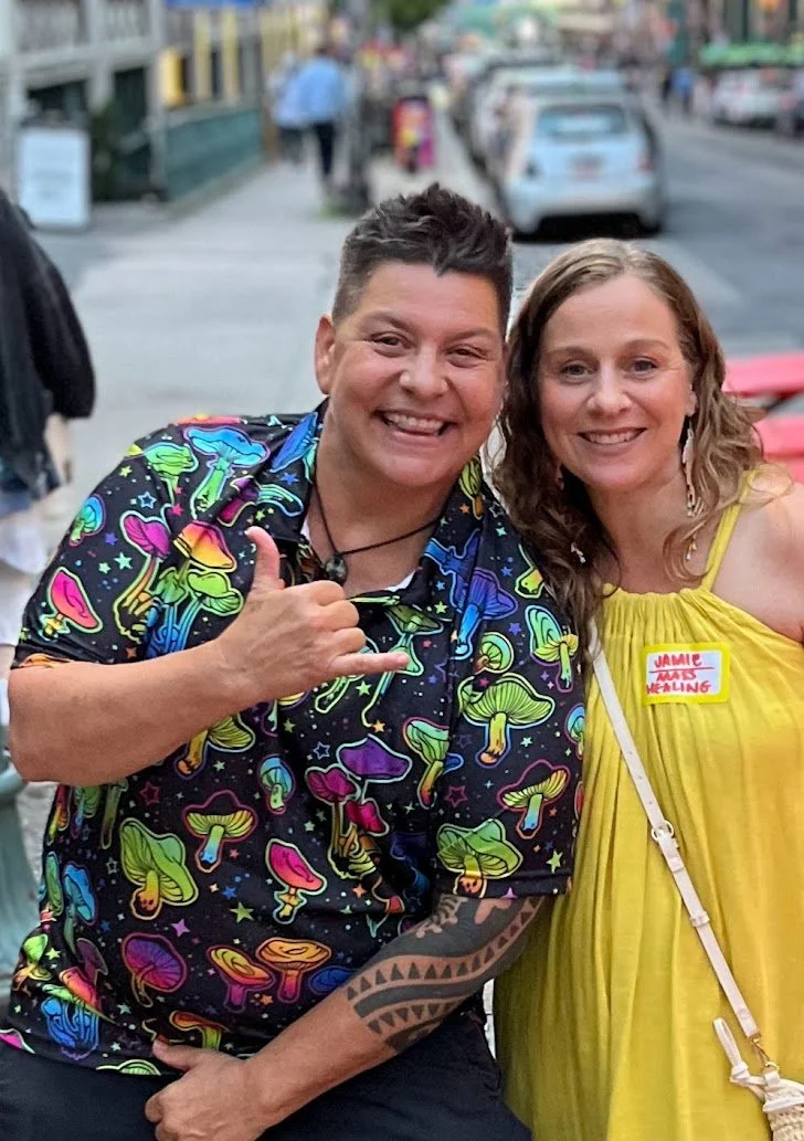 A man and woman smiling on a city street. The man is wearing a colorful mushroom-patterned shirt. The woman is wearing a yellow dress with a name tag that reads "Jamie has healing." There are cars and pedestrians in the background.