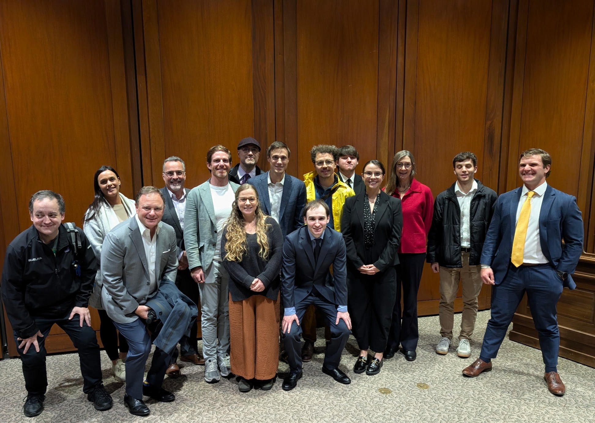 A group of diverse people posing together indoors in front of a brown wooden wall, some smiling and some with neutral expressions.