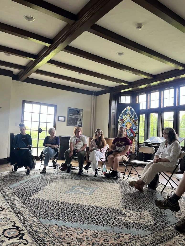 Six people sitting in a semi-circle in a room with large windows, a stained glass window, and a patterned area rug.