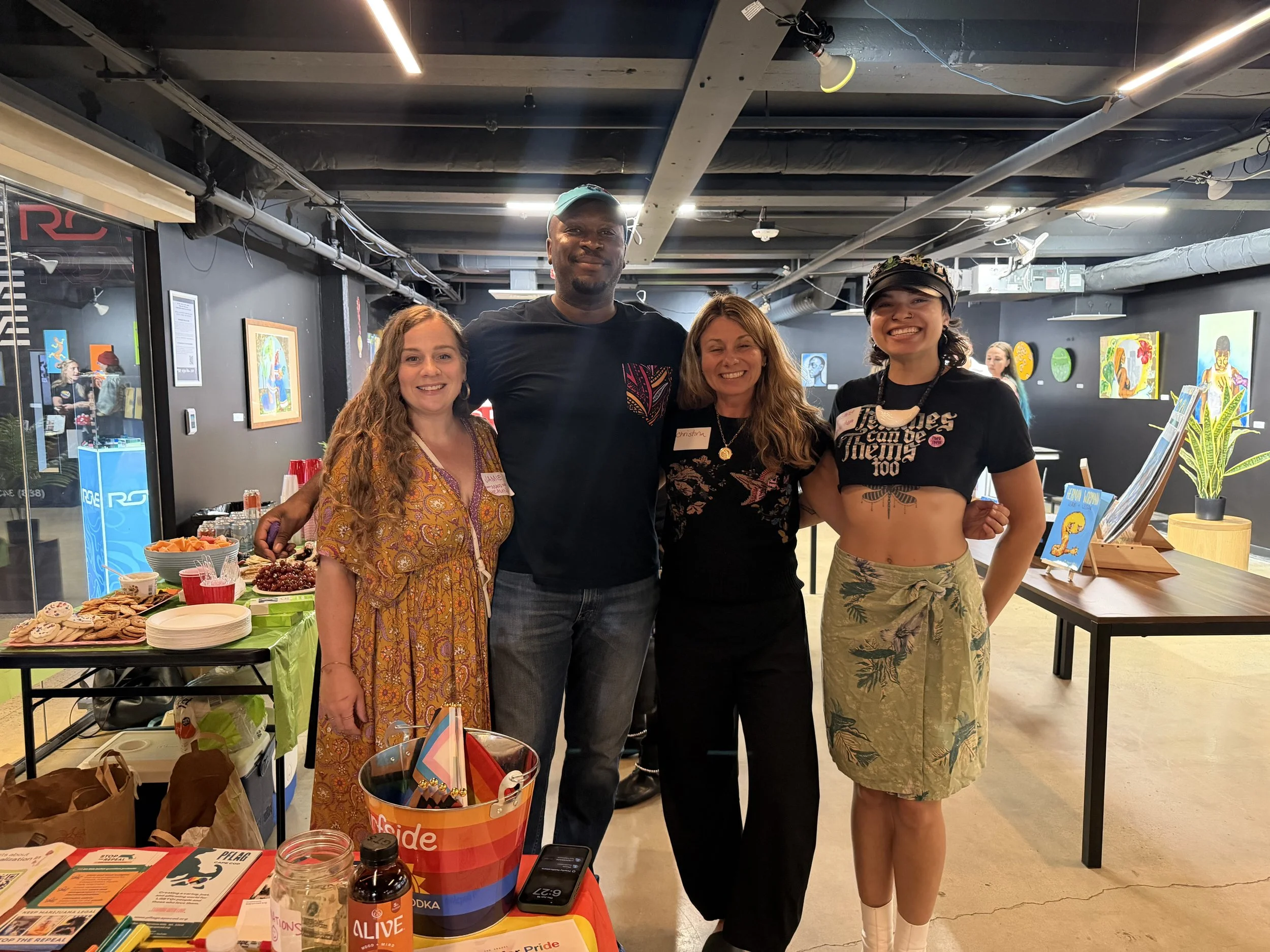Group of four people standing indoors at a social event, smiling, with tables and colorful decorations around them.