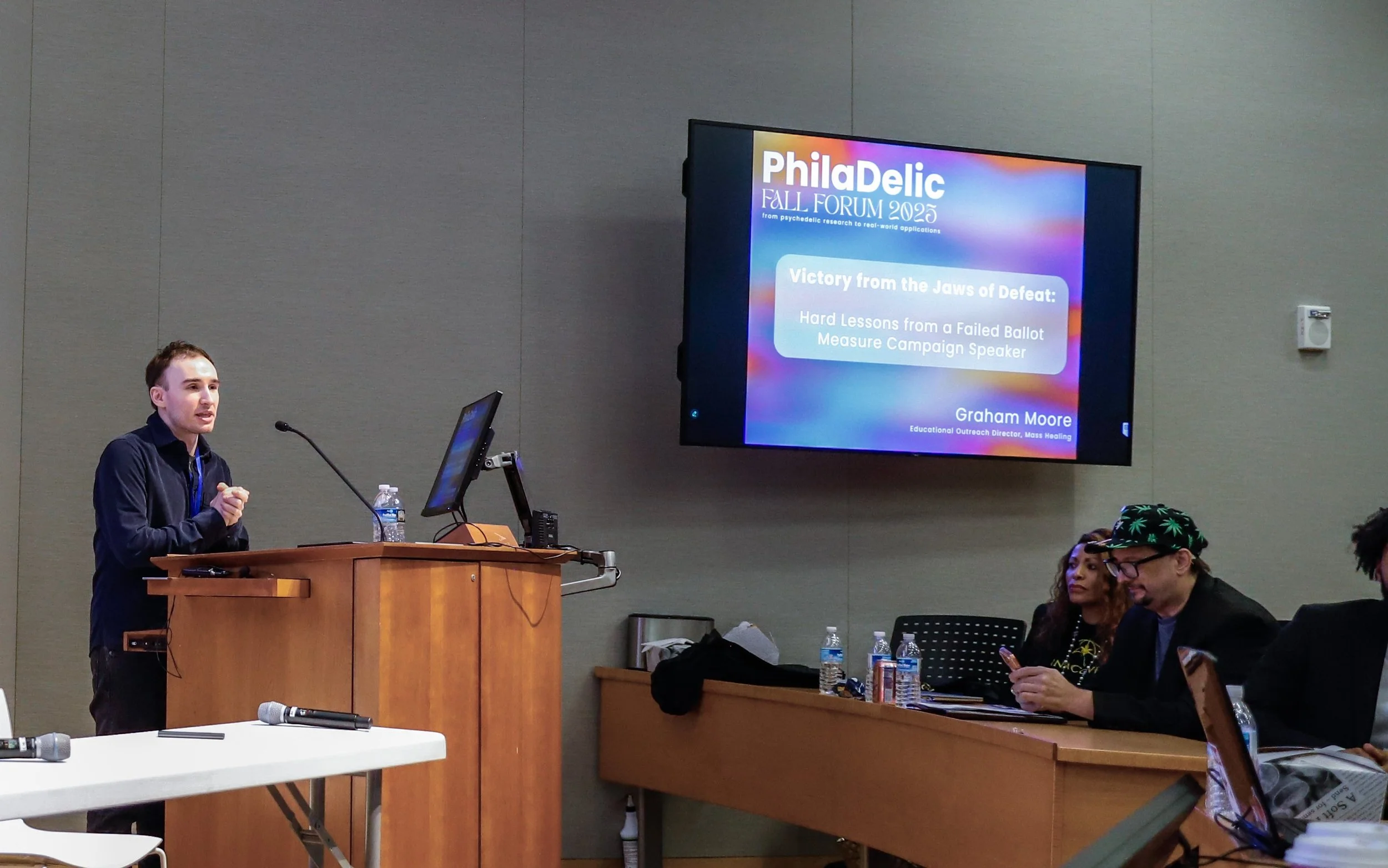 A man standing at a podium giving a presentation at the Philadelic Fall Forum 2025. Behind him is a large screen displaying the event's name and topic, with three people seated at a long table to the right, and attendees in the foreground.