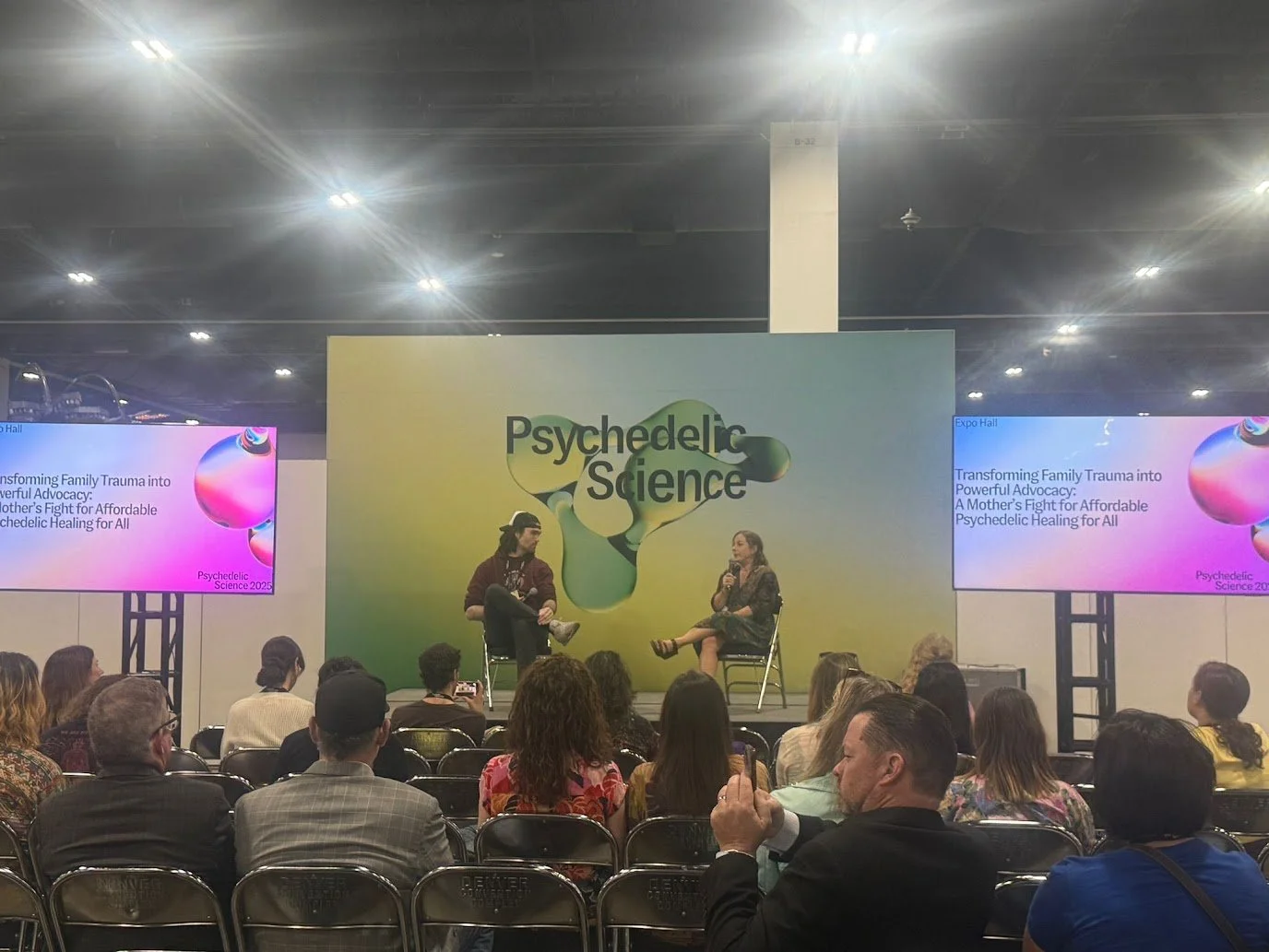 Psychedelic Science '25 conference. A stage featuring two women seated and talking, behind a large sign that says 'Psychedelic Science.' Audience members are sitting in rows facing the stage. Two screens display the event topic: 'Transforming Fa