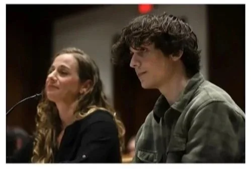 A woman with wavy hair smiling and a young man with curly hair sitting next to her in a room with dark background.