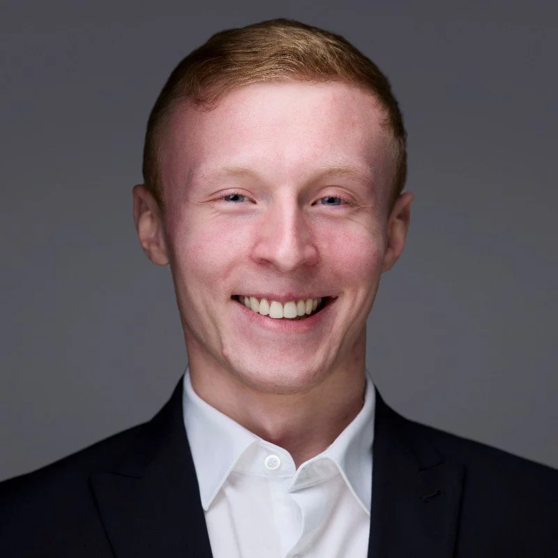 Headshot of a young man with short red hair, blue eyes, and a wide smile, wearing a black suit jacket and white shirt, against gray background.