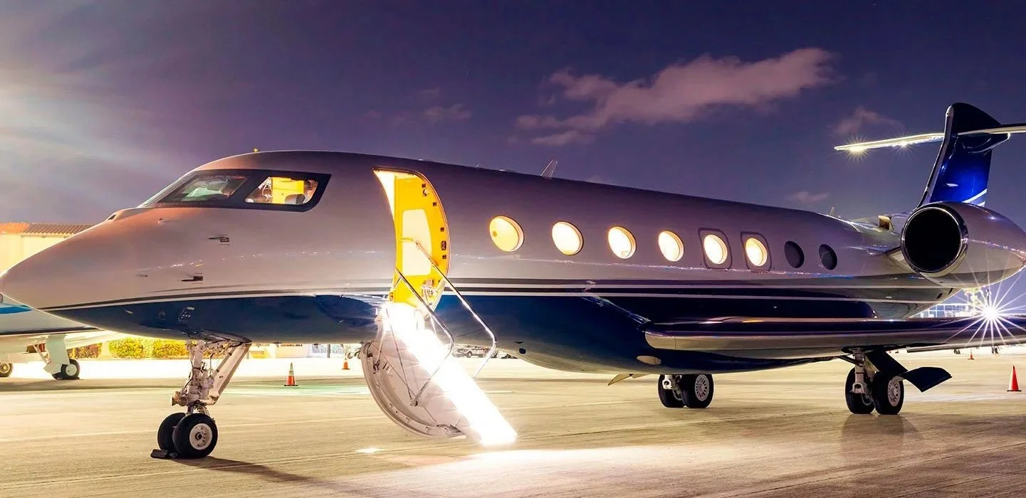 A luxury private jet, Gulfstream G650 is parked on an airport tarmac at night, with its door open and stairs extended. The jet is sleek and modern with a dark blue and silver exterior, and the sky is dark with a few clouds.
