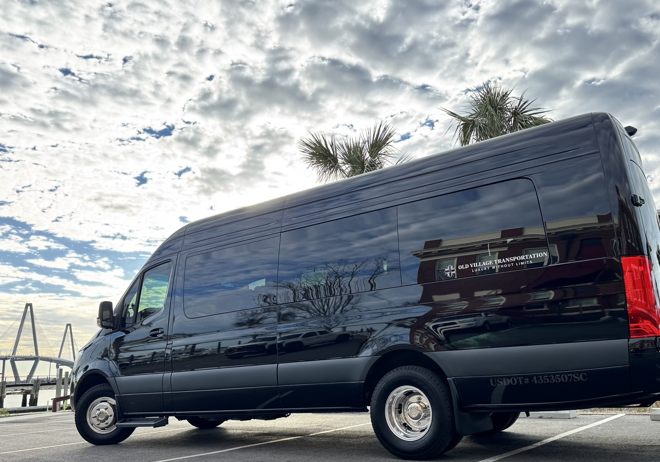 Our Mercedes Benz Executive Sprinter 3500 XD parked at a Private Marina on Charleston Harbor with palm trees and a the Ravenel bridge in the background, reflecting the cloudy sky and surroundings on its shiny surface.