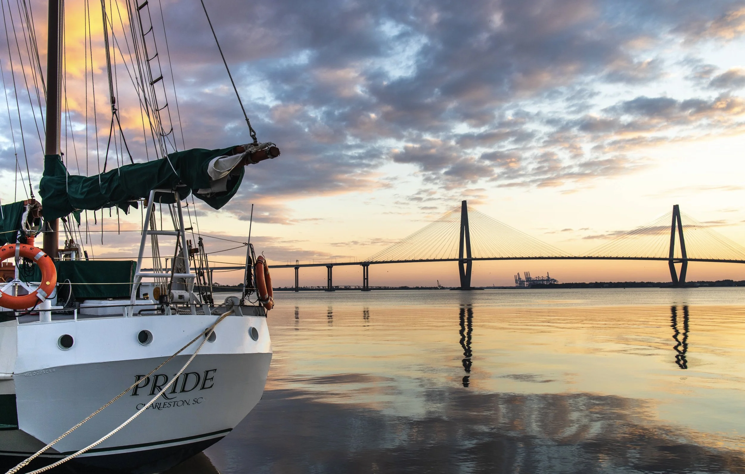 A sailboat named 'PRIDE' docked on Charleston Harbor during sunset with Ravenel bridge and a partly cloudy sky in the background.
