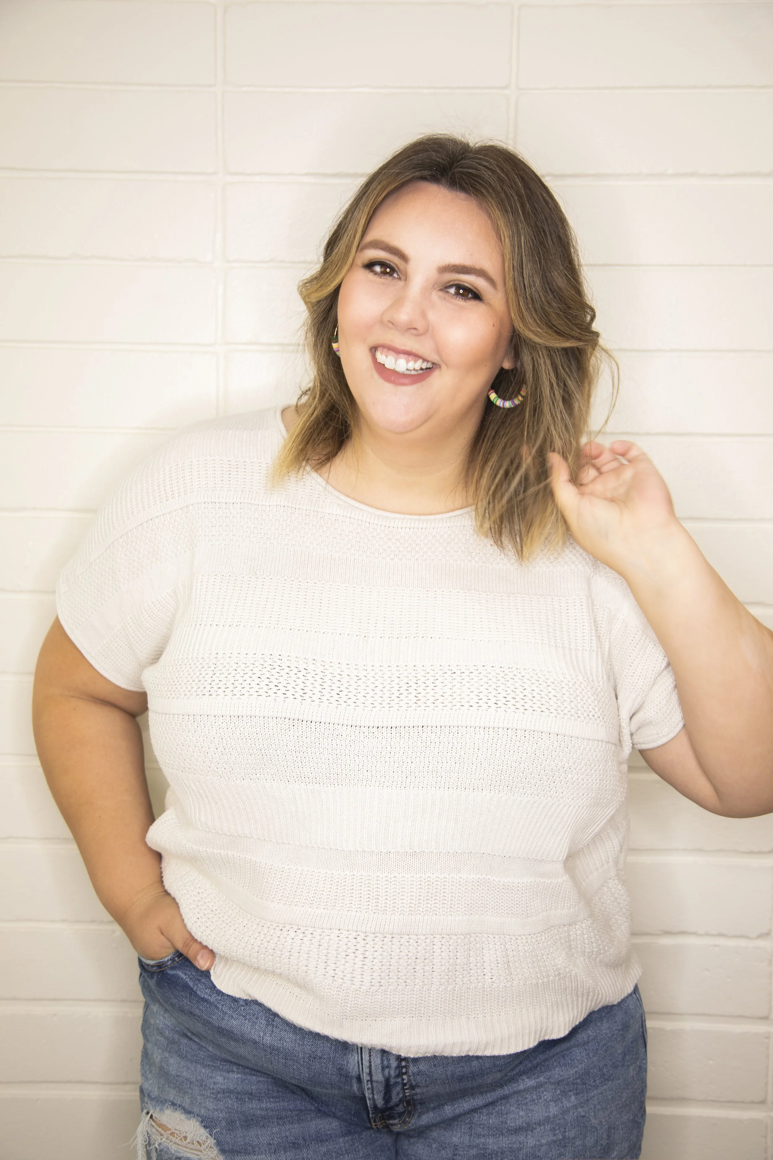 A woman, who is a certified birth and postpartum doula with shoulder-length hair smiling, wearing a white knit shirt and blue jeans, standing in front of a white brick wall.