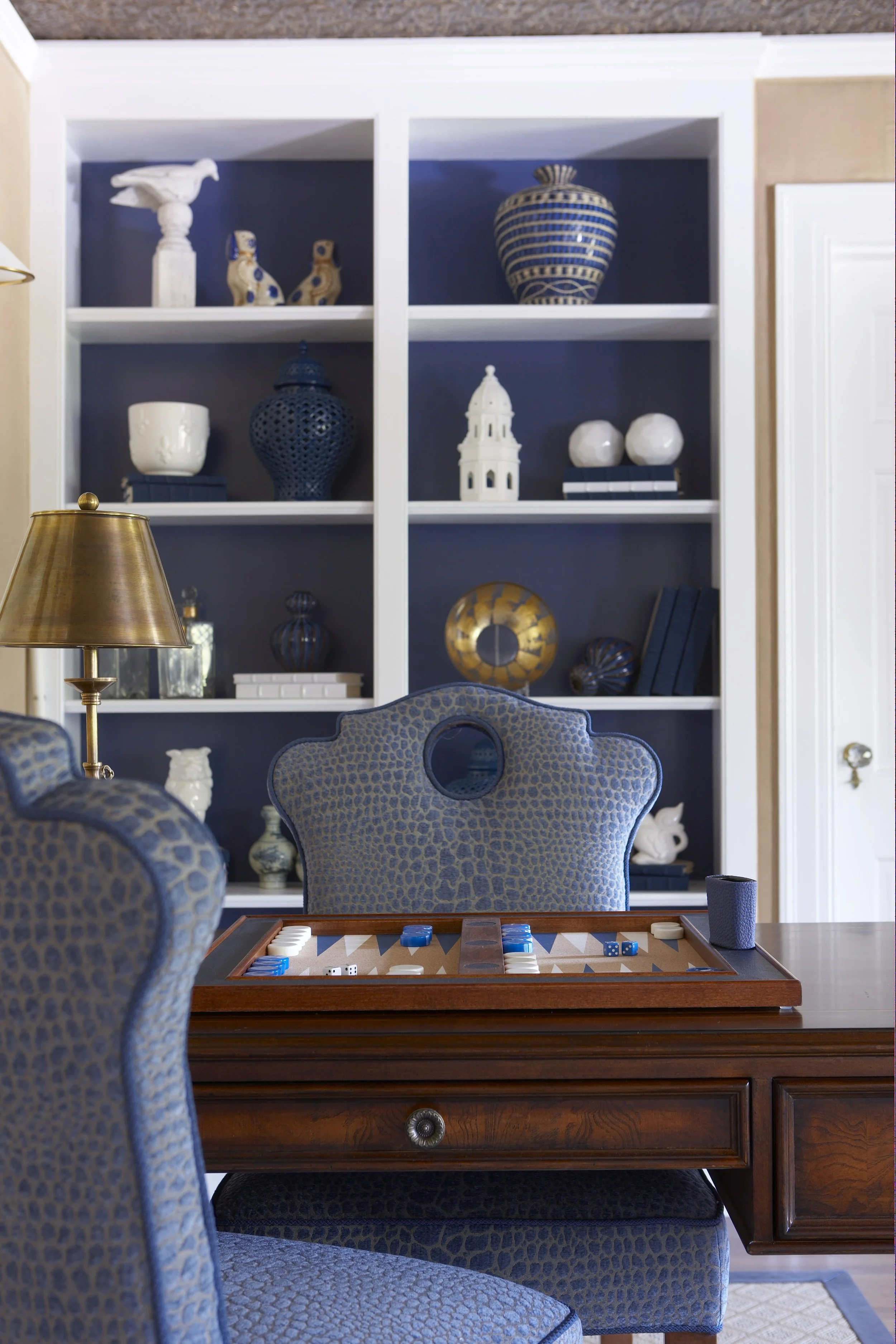 Room interior with blue upholstered chairs, a wooden table with a backgammon set, and built-in shelves displaying decorative vases and ornaments.