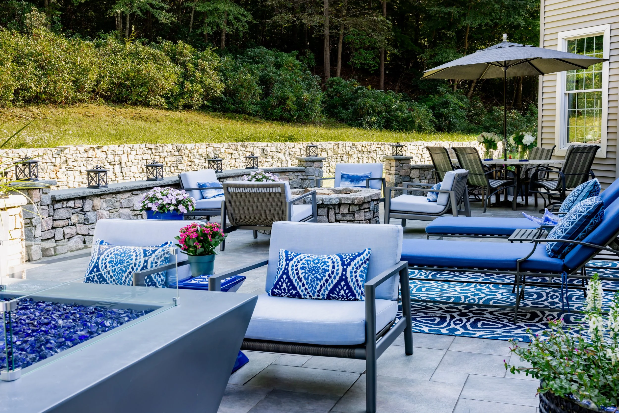 Outdoor patio with blue and white furniture, decorative pillows, stone fire pit, and an umbrella. Surrounded by a stone wall, potted flowers, and greenery.