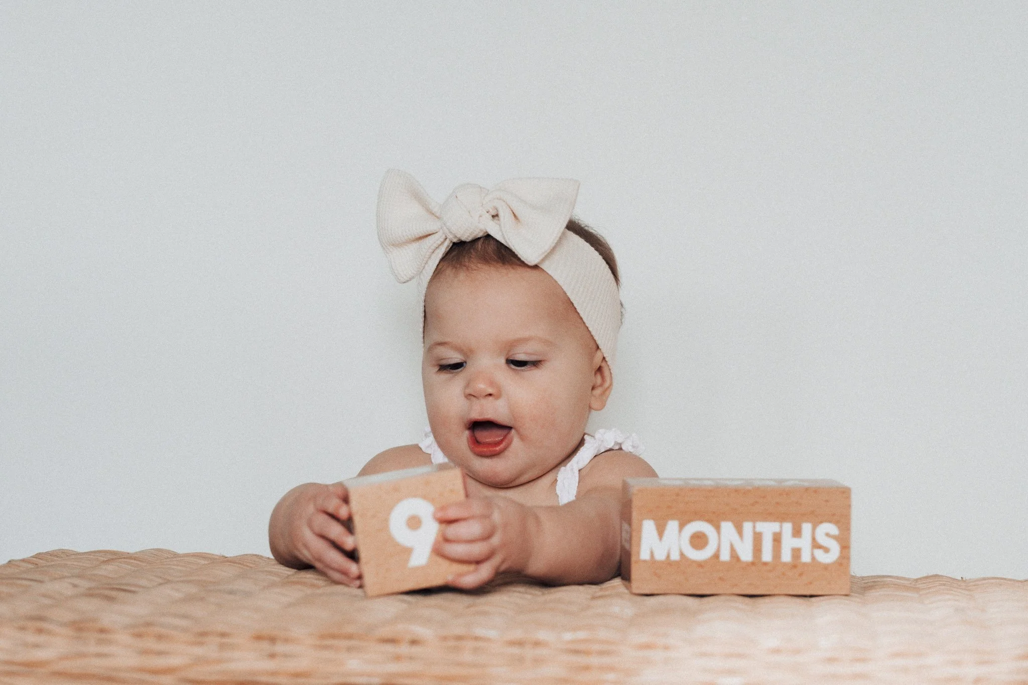 Baby girl wearing a large beige bow headband, sitting at a table, holding a wooden block with the number 9, with another block labeled 'MONTHS' in front of her.