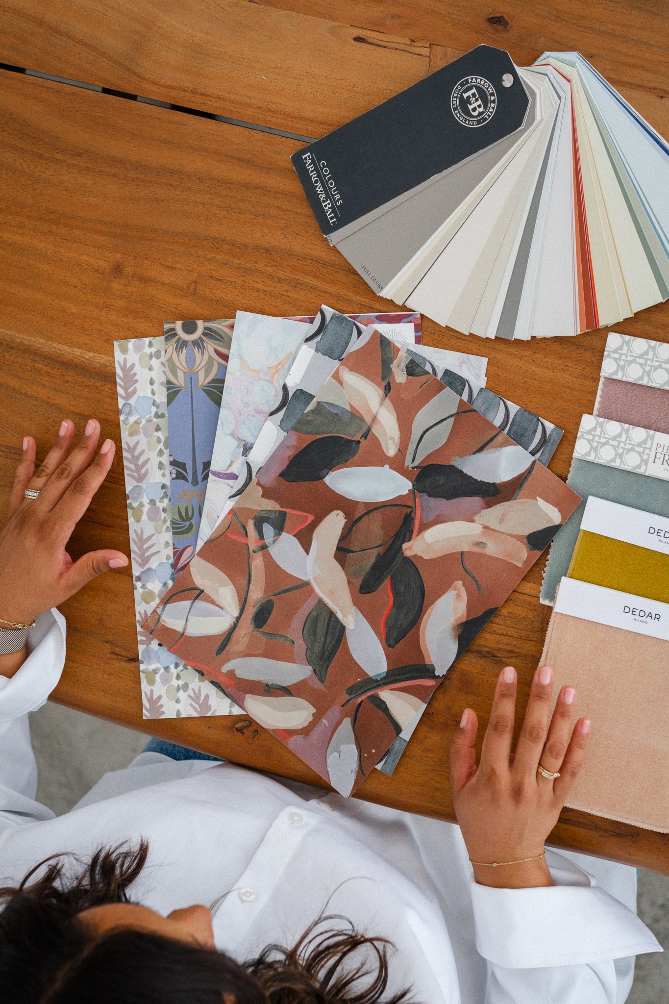 Person sitting at a wooden table with various fabric samples, color swatches, and paint color cards, including a fan deck of paint chips. The person is wearing a white shirt and has their hands resting on the table.