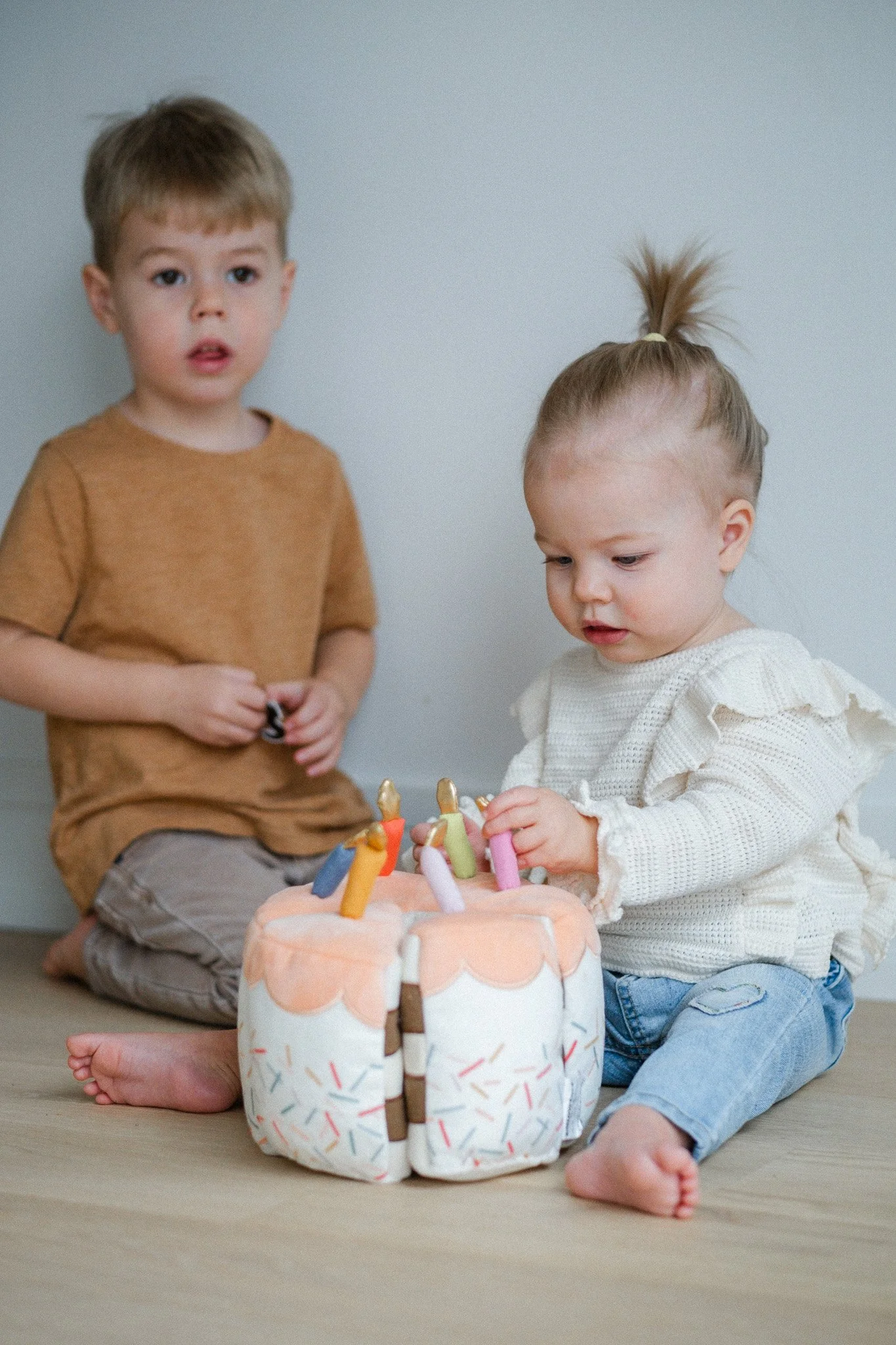 Two young children celebrating a birthday with a cake featuring lit candles, sitting on a wooden floor.