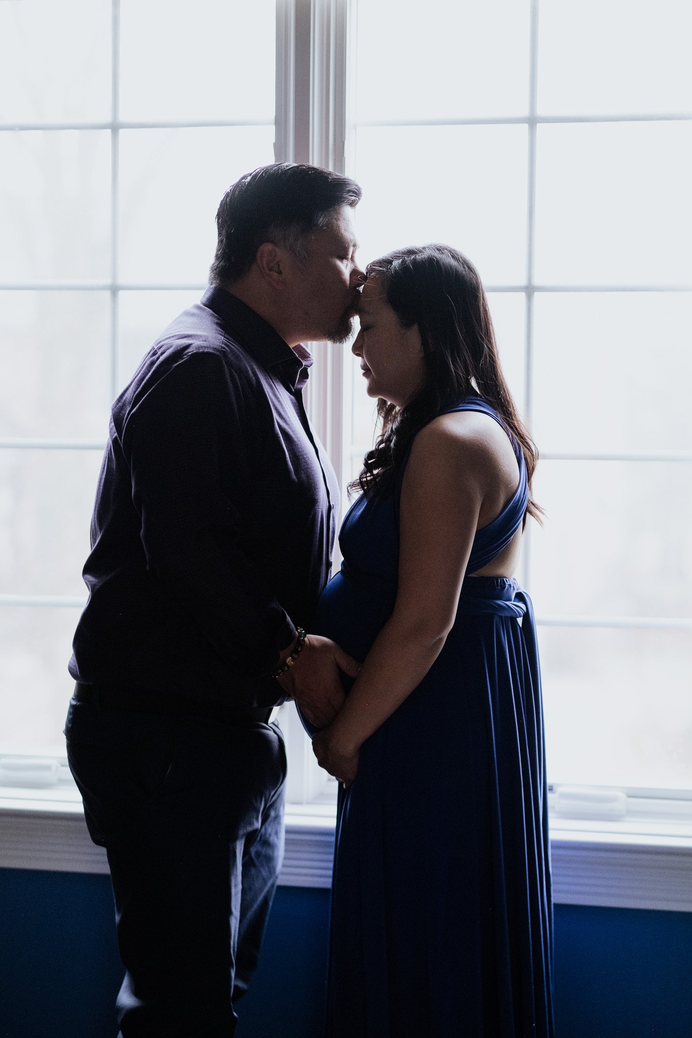 A couple standing close together near a large window, with the man gently kissing the woman on the forehead. The woman has her eyes closed and is holding her pregnant belly. The scene appears intimate and tender, set indoors with backlighting from the window.