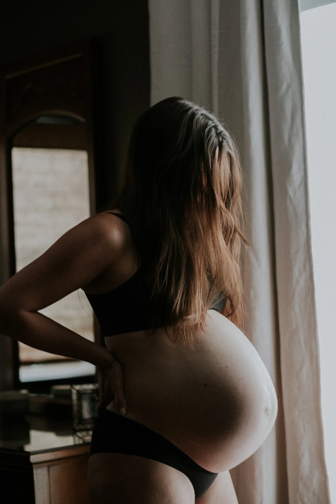 Pregnant woman standing indoors near a window, with her hand on her back, wearing black underwear and a black top, with her head slightly bowed and face partially covered by her hair.