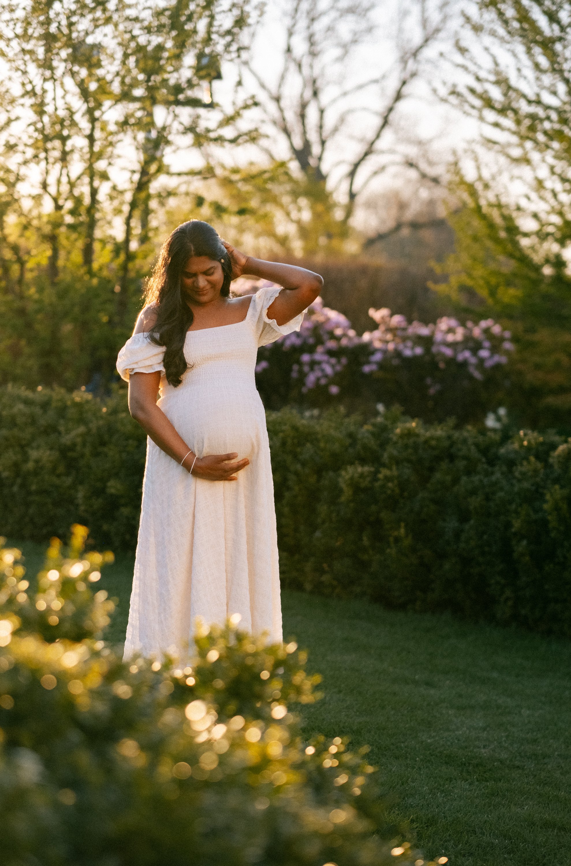 A pregnant woman in a white dress gently holding her belly with one hand and touching her head with the other, standing outdoors in a garden during sunset.