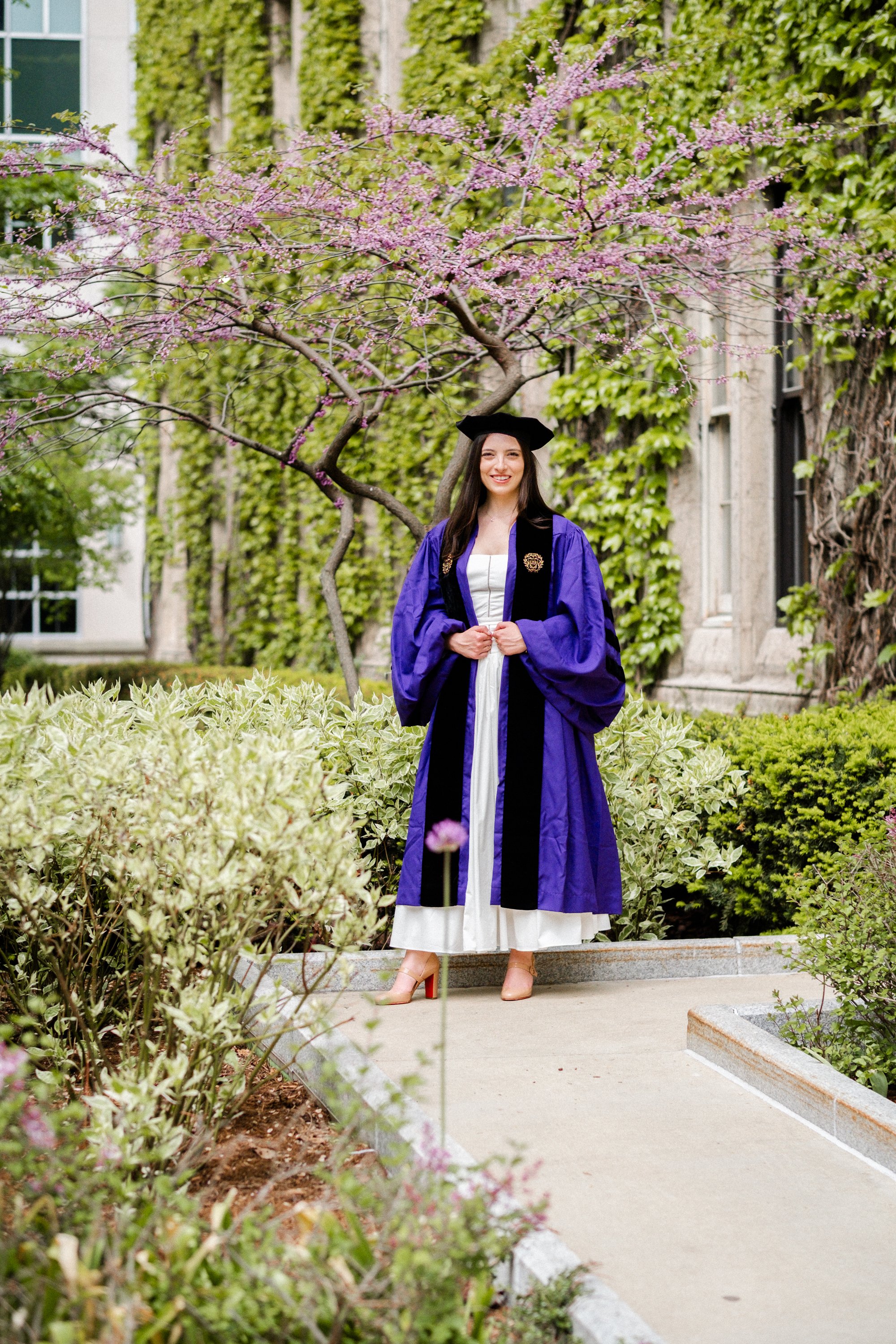 A woman in a purple graduation gown and cap standing outdoors next to a pink flowering tree, smiling.