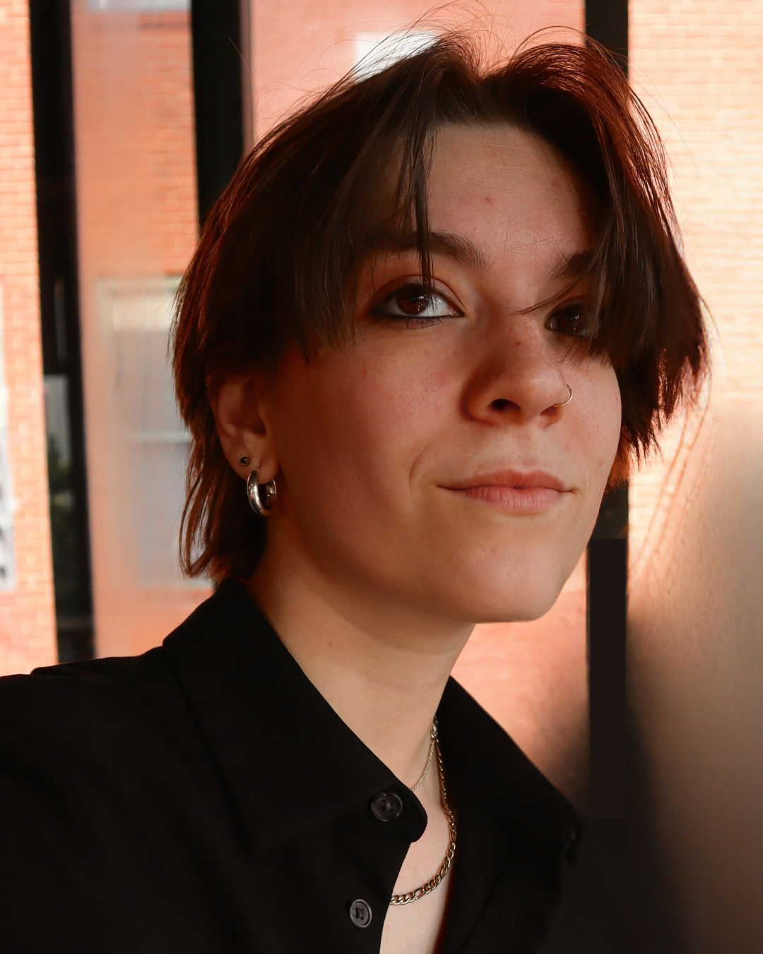 A young person with short brown hair and multiple earrings wearing a black shirt, standing indoors against a wall, with windows and a brick building in the background.