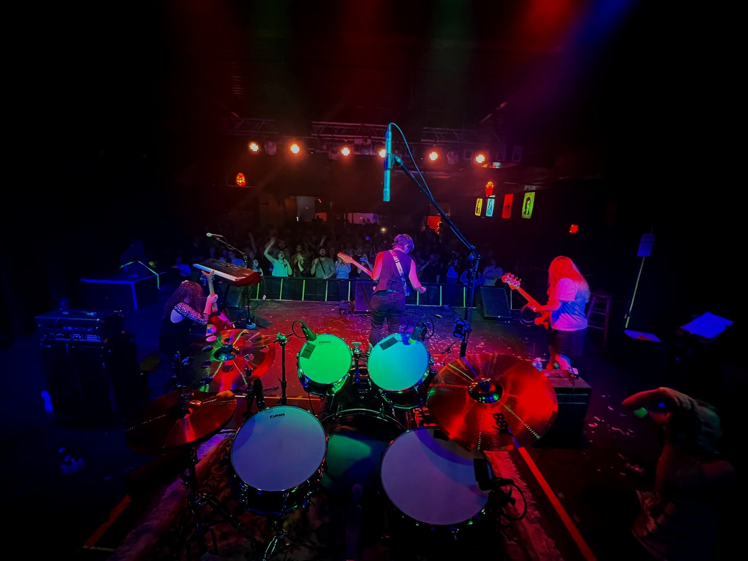 A band performing live on stage with a drum set, electric guitars, and a keyboard, under colorful stage lights in front of a crowded audience.