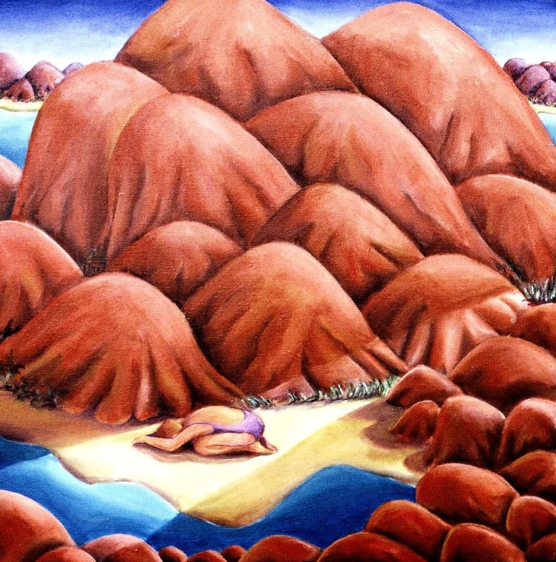 A person lying on a sandy beach near a body of water, surrounded by large reddish-brown rocks and mountains in the background.