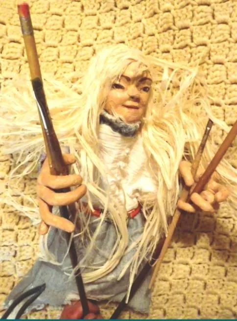 A puppet with long rafia hair, jean skirt, silk top, and paint brushes and pencil sticks in her large hands.