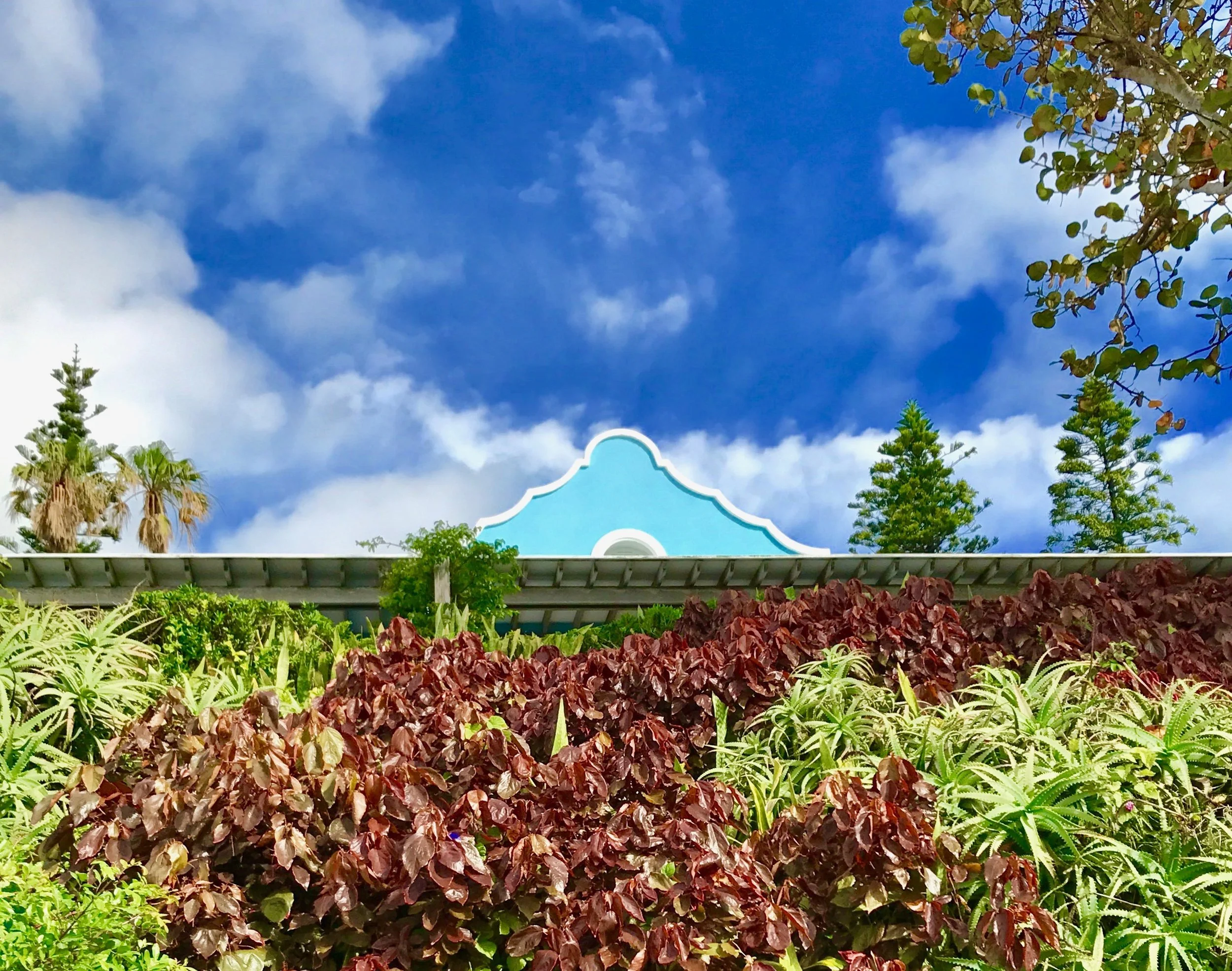 Blue sky with white clouds and a building with a blue facade and decorative roof edge, surrounded by trees and lush greenery.